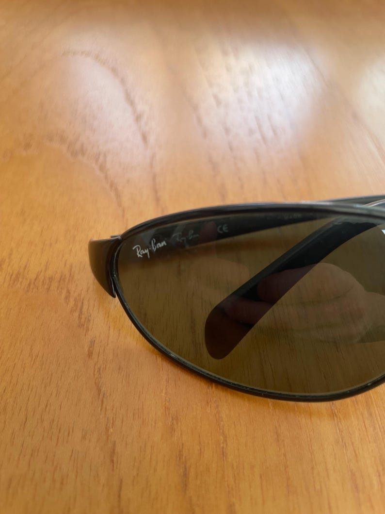 Puede incluir: Primer plano de gafas de sol Ray-Ban con lentes oscuras y montura negra. El logotipo de Ray-Ban es visible en la lente izquierda. Las gafas de sol est&aacute;n sobre una superficie de madera.