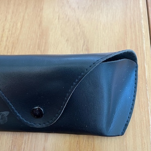 Puede incluir: Un estuche negro para gafas de sol, de aspecto similar al cuero, con cierre de bot&oacute;n. El estuche rectangular tiene una parte superior curvada y est&aacute; colocado sobre una superficie de madera de color marr&oacute;n claro. El estuche parece estar vac&iacute;o.