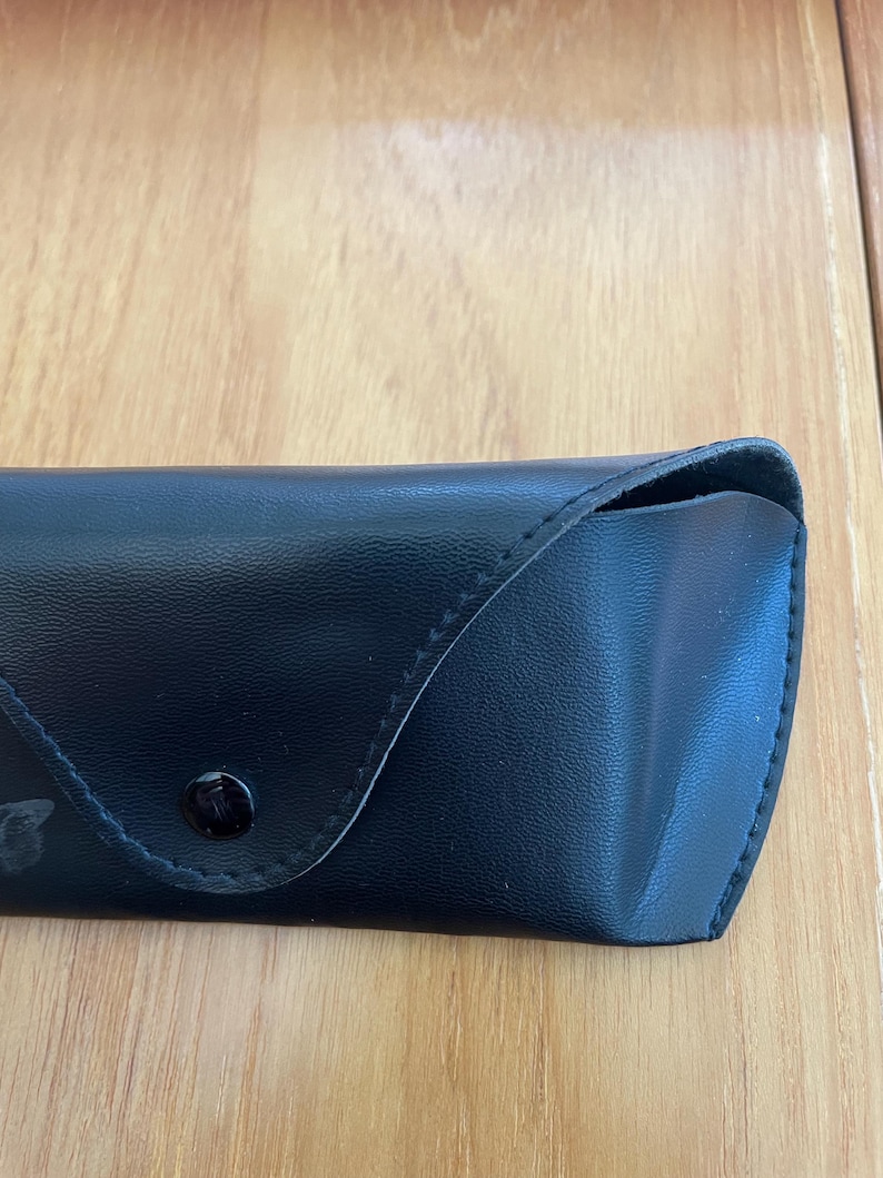 Puede incluir: Un estuche negro para gafas de sol, de aspecto similar al cuero, con cierre de bot&oacute;n. El estuche rectangular tiene una parte superior curvada y est&aacute; colocado sobre una superficie de madera de color marr&oacute;n claro. El estuche parece estar vac&iacute;o.
