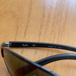 Puede incluir: Gafas de sol negras con lentes tintadas. El logotipo de Ray-Ban es visible en la patilla, junto con el texto "CE" y "MADE IN ITALY". Las gafas de sol est&aacute;n sobre una superficie de madera.