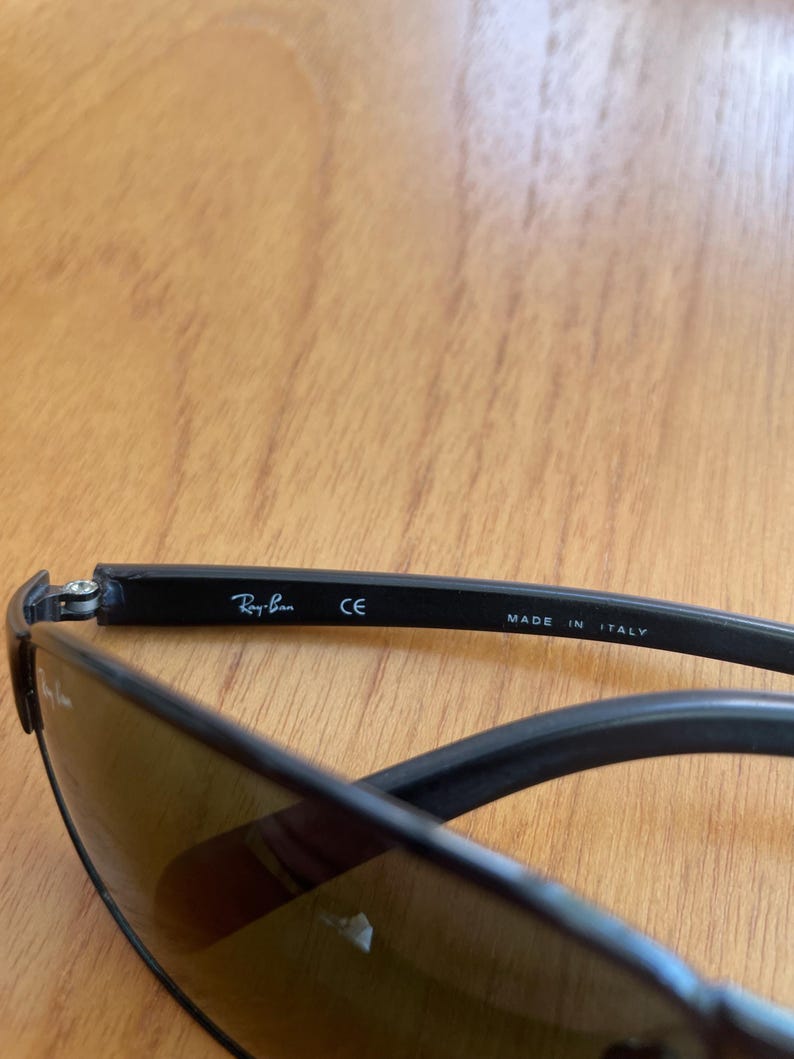 Puede incluir: Gafas de sol negras con lentes tintadas. El logotipo de Ray-Ban es visible en la patilla, junto con el texto "CE" y "MADE IN ITALY". Las gafas de sol est&aacute;n sobre una superficie de madera.