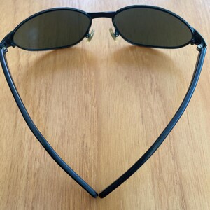 Puede incluir: Gafas de sol negras con lentes ovaladas y una montura negra fina. Las gafas de sol tienen un dise&ntilde;o cl&aacute;sico con un aspecto elegante y moderno. Las lentes son de color marr&oacute;n oscuro y las patillas se curvan hacia atr&aacute;s para un ajuste c&oacute;modo.