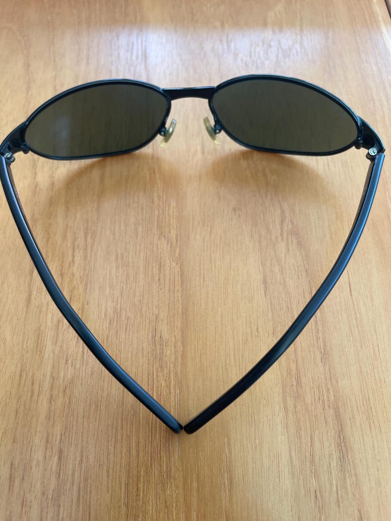 Puede incluir: Gafas de sol negras con lentes ovaladas y una montura negra fina. Las gafas de sol tienen un dise&ntilde;o cl&aacute;sico con un aspecto elegante y moderno. Las lentes son de color marr&oacute;n oscuro y las patillas se curvan hacia atr&aacute;s para un ajuste c&oacute;modo.