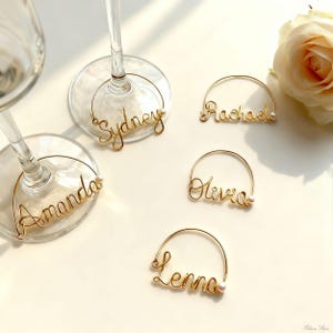 Può includere: Ciondoli per calici in filo d'oro con i nomi "Sydney", "Amanda", "Rachael", "Olivia" e "Lenna". Ogni ciondolo ha un piccolo accento di perla. Una rosa bianca è sullo sfondo.