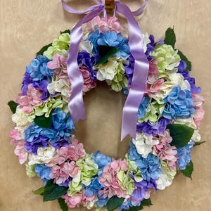 Puede incluir: Una colorida corona floral con flores de hortensia en tonos rosa, azul, morado, verde y blanco. La corona está adornada con hojas verdes y una cinta lavanda para colgar. Un acento decorativo para cualquier puerta.
