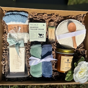 Puede incluir: Una caja de regalo llena de artículos de cuidado personal. Incluye calcetines azules, crema de sebo batida, una caja de cerillas, una cuchara de madera, una vela de vainilla, jabón de leche de cabra y dos toallas pequeñas. También incluye una flor blanca y vegetación.