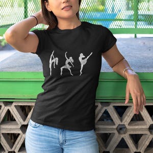 May include: Black t-shirt with a white graphic of three ballet dancers in different poses. The shirt is made of a soft material and is paired with light blue jeans. The model is wearing a black bracelet and a gold bracelet.
