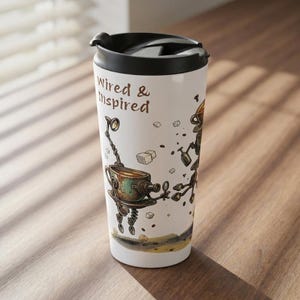 Könnte beinhalten: Weißer Reisebecher mit schwarzem Deckel, mit dem Text "Wired & Inspired" und einer skurrilen Illustration eines Roboters, der eine Teetasse hält. Das Design umfasst Braun-, Kupfer- und Beigetöne.
