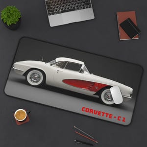 Peut inclure: Tapis de souris Corvette C1 blanc et rouge. Le tapis de souris présente une Corvette blanche classique avec des accents rouges et le texte "CORVETTE - C1" en rouge. Le tapis de forme rectangulaire est posé sur une surface gris foncé.