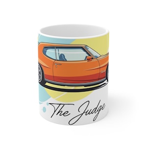 May include: White ceramic mug featuring an orange cartoon car with black wheels. The car is set against a yellow and blue background. The words "The Judge" are written in black script on the mug.