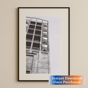 Puede incluir: Fotografía en blanco y negro de un edificio alto de hormigón con grandes ventanales geométricos. El edificio está inclinado hacia el espectador, y el cielo es visible en el fondo.