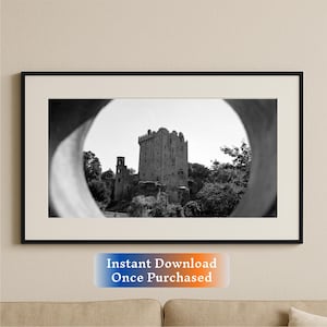 May include: Black and white photograph of a stone castle framed in a black frame. The castle is seen through a circular opening in a stone wall. The castle is surrounded by trees.