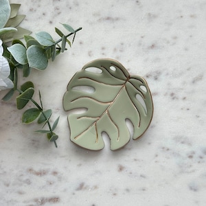 May include: A light green ceramic dish shaped like a monstera leaf with gold leaf detailing. The dish is perfect for holding jewelry, keys, or other small items.