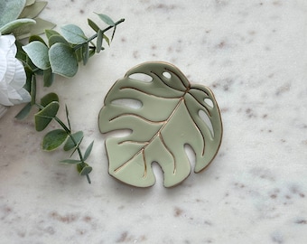 May include: A light green ceramic dish shaped like a monstera leaf with gold leaf detailing. The dish is perfect for holding jewelry, keys, or other small items.