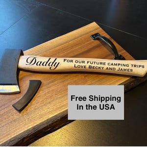 May include: Axe with a wooden handle and a dark metal blade, engraved with "Daddy" and a message about camping trips. The axe is placed on a wooden cutting board with a black handle.