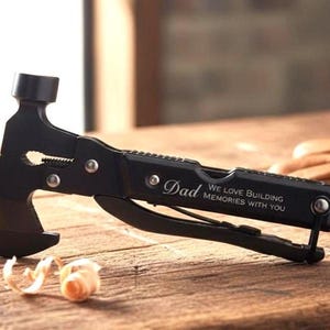 May include: A black multi-tool with a hammer, axe, and wrench. The tool has the words "Dad" and "We love building memories with you" engraved on it. The tool is on a wooden surface.