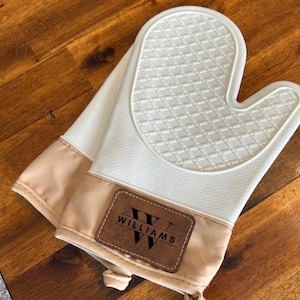 May include: A white silicone oven mitt with a textured diamond pattern on the palm area. The cuff is made of beige fabric and features a rectangular brown leather patch with the black text WILLIAMS and a large letter W printed on it.