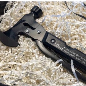 May include: A black multi-tool hammer with a hatchet head, hammer, and folding tools. The handle is engraved with the word "Dad" and the phrase "We love building memories with you."