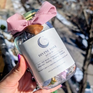 May include: A clear glass jar labeled "DIVINE LOVE Spell Jar Kit" with a pink ribbon and wooden accent. The label lists "Love, Self-Worth, Compassion, Passion, Attraction, Healing, Harmony, Connection." The jar contains dried flowers and herbs.