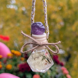 May include: A small, clear glass bottle with a diamond-shaped design, filled with a light-colored liquid and botanical elements. The bottle is topped with a wooden stopper, a purple gemstone, and a twine bow. It hangs from a braided cord.