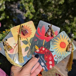 May include: A collection of four decorative bookmarks with floral and sun illustrations. Each bookmark has a small glass vial filled with dried flowers and a gold tassel. The text "Sunrise brings possibilities" is visible on one bookmark.