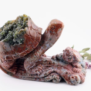May include: A carved stone turtle sculpture in shades of red, brown, and green. The turtle has a detailed shell and is accompanied by a smaller turtle. The sculpture is set on a base with wave-like patterns.