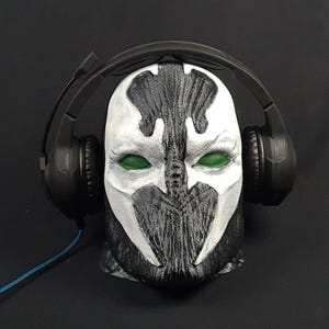 May include: A detailed bust of the Spawn character with a black and white mask and green eyes, wearing a black headset. The bust is set against a dark background, with a blue cable visible.