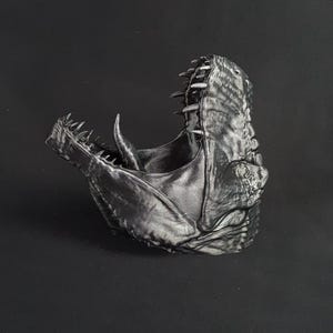 Puede incluir: Una máscara detallada con la boca abierta en tonos grises, que se asemeja a una cabeza de dinosaurio o dragón. La máscara presenta dientes afilados y piel texturizada, creando una apariencia realista e intimidante. La máscara es probablemente para disfraz o exhibición.