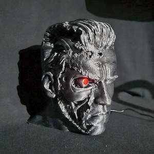 Può includere: Un busto nero scolpito della testa di un cyborg con un occhio rosso brillante. La testa è dettagliata con componenti meccanici esposti e danni da battaglia. Il busto è posto su uno sfondo scuro, evidenziando il design intricato.