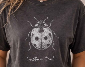 May include: A charcoal gray short-sleeve t-shirt featuring a white distressed graphic of a ladybug centered on the chest. Below the ladybug, the words Custom text are printed in a white script font. The shirt has a crew neckline.
