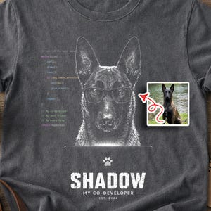 May include: Dark gray t-shirt featuring a dog wearing glasses, with the text "SHADOW MY CO-DEVELOPER EST. 2024". A small image of the dog is on the shirt. A coffee cup, sunglasses, and a notepad are on a wooden surface.