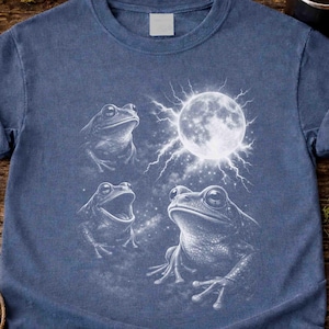 May include: Denim blue t-shirt featuring a detailed white illustration of three frogs and a full moon with radiating lines. The frogs are in different poses, with one open-mouthed. The word "Denim" is printed on the bottom right.