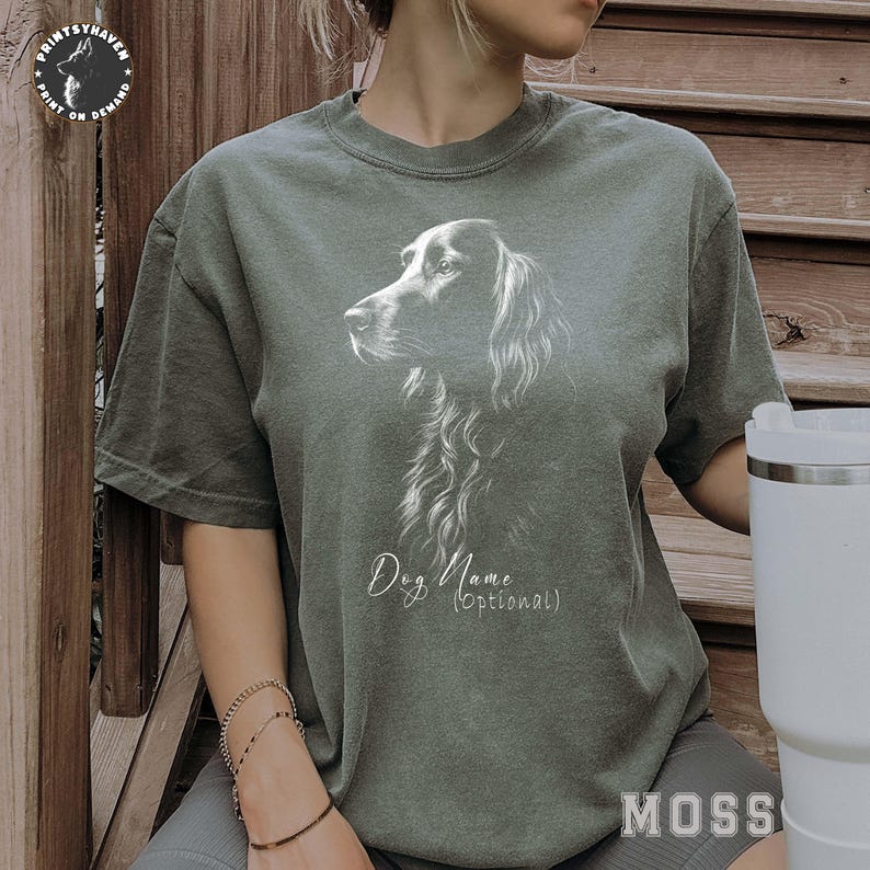 May include: A faded green t-shirt featuring a detailed white illustration of a dog's head and chest. The text "Dog Name (Optional)" is below the dog image. The shirt is part of the "MOSS" collection.