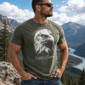 May include: An olive green short-sleeved t-shirt featuring a white graphic of a bald eagle head in profile. Below the eagle, the text Protector of Freedom is printed in a white script font. The shirt has a crew neckline and a standard fit.