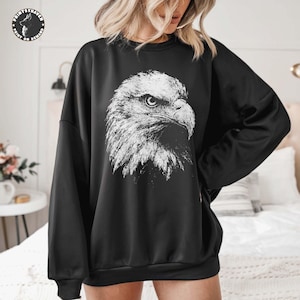 May include: A black long-sleeve sweatshirt featuring a large, detailed white graphic print of a bald eagle head in profile on the centre chest. The eagle design displays intricate feather textures and a sharp, focused gaze.
