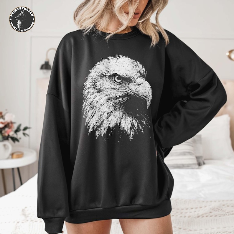 May include: A black long-sleeve sweatshirt featuring a large, detailed white graphic print of a bald eagle head in profile on the centre chest. The eagle design displays intricate feather textures and a sharp, focused gaze.