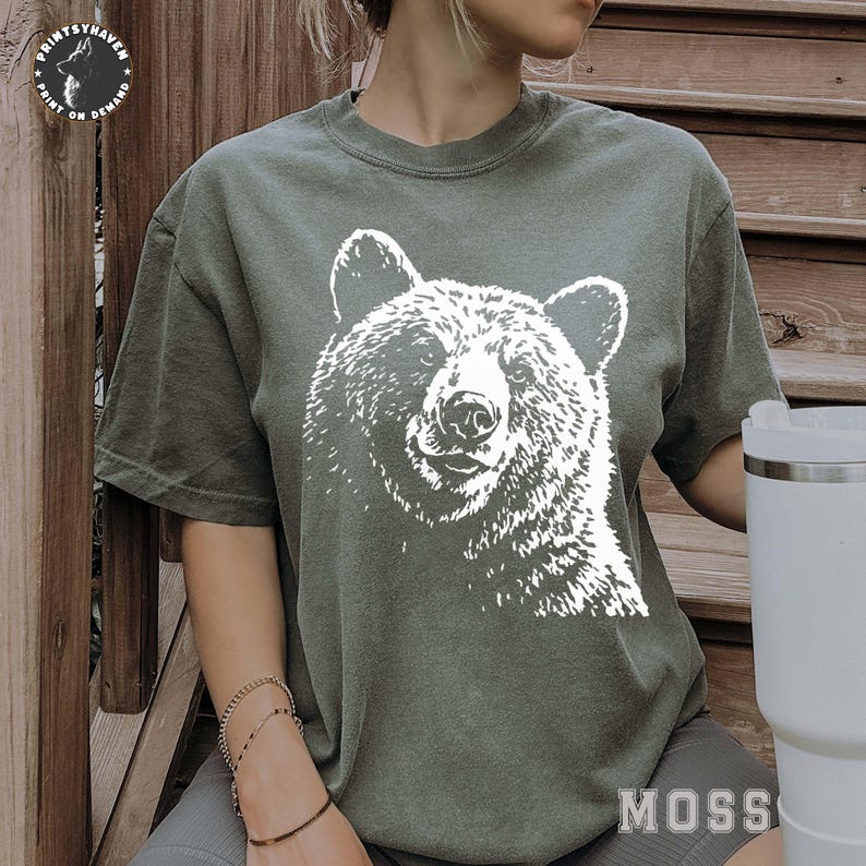 Puede incluir: Camiseta verde con un gr&aacute;fico blanco de la cara de un oso. La camiseta tiene mangas cortas y cuello redondo. La palabra "MOSS" est&aacute; impresa en blanco en la parte inferior derecha.