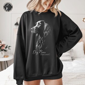 May include: Black sweatshirt featuring a detailed grayscale illustration of a dog's profile. The design includes the text "Dog Name (optional)" in a script font. The sweatshirt has a crew neck and long sleeves.