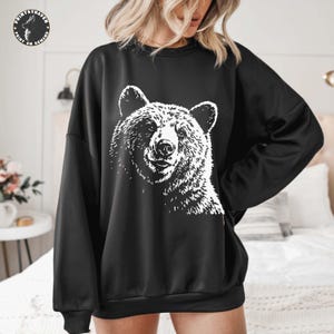 Puede incluir: Sudadera negra con un gr&aacute;fico blanco de la cara de un oso. El pelaje del oso est&aacute; detallado con l&iacute;neas texturizadas, y el dise&ntilde;o est&aacute; centrado en la parte delantera de la parte superior de manga larga. La sudadera tiene cuello redondo y un corte holgado.