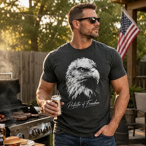 May include: A charcoal grey short-sleeve t-shirt featuring a white graphic illustration of a bald eagle head and the text "Protector of Freedom" printed in a cursive script font across the chest area of the garment.