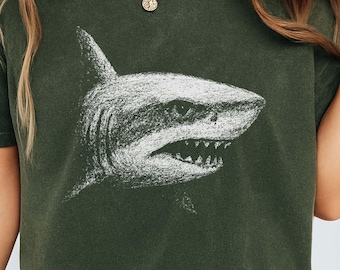May include: Dark green t-shirt featuring a white shark illustration. The shark is depicted in a detailed, sketched style, with visible teeth and fins. The word "HEMP" is printed in a bold font at the bottom of the shirt.