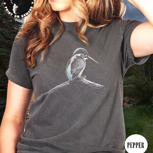 May include: Dark grey t-shirt featuring a detailed illustration of a kingfisher bird perched on a branch. The shirt has short sleeves and a crew neck. The text "Printshaven" and "Print on Demand" is printed on the top left of the shirt. The word "Pepper" is printed on the bottom right.