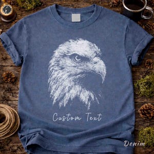 May include: A blue short-sleeved t-shirt featuring a white graphic illustration of a bald eagle head in profile. The design includes the text "Custom Text" printed in a white script font below the eagle graphic on the front of the garment.