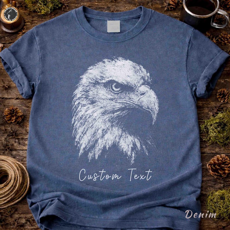 May include: A blue short-sleeved t-shirt featuring a white graphic illustration of a bald eagle head in profile. The design includes the text "Custom Text" printed in a white script font below the eagle graphic on the front of the garment.