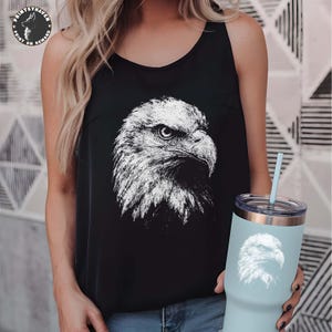 May include: A black sleeveless tank top featuring a white graphic print of a bald eagle head on the front. The tank top is paired with a light blue insulated tumbler that also displays a matching white bald eagle head graphic and a light blue straw.