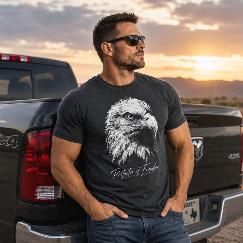 May include: A charcoal grey short-sleeve t-shirt featuring a white graphic of a bald eagle head and the text Protector of Freedom printed in a script font. The shirt has a crew neckline and a standard fit, showcasing a detailed wildlife illustration.