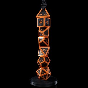 Op de afbeelding: Een stapel zwarte en oranje veelvlakkige dobbelstenen, waaronder een D4, D6, D8, D10, D12 en D20, met zwarte cijfers. De dobbelstenen staan op een zwarte basis, met een zwart lint aan de bovenkant. De achtergrond is zwart.