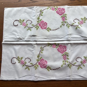 May include: Two white rectangular pillowcases featuring a cross-stitched floral pattern of pink roses with green leaves and brown stems arranged in a circular wreath design. The bottom edges of the pillowcases have a scalloped hem finish.