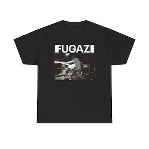 Puede incluir: Camiseta negra con el nombre de la banda "Fugazi" en letras blancas en bloque sobre una imagen en blanco y negro de un miembro de la banda tocando el bajo en el escenario. La camiseta es de cuello redondo.