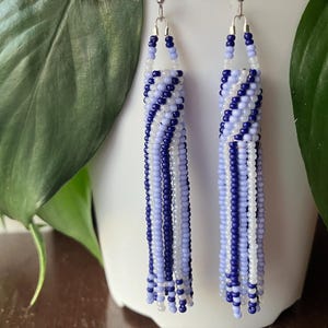 May include: A pair of long, beaded earrings with a blue and white color scheme. The earrings are made of small, round beads that are strung together in a pattern. The earrings are hanging from silver hooks.